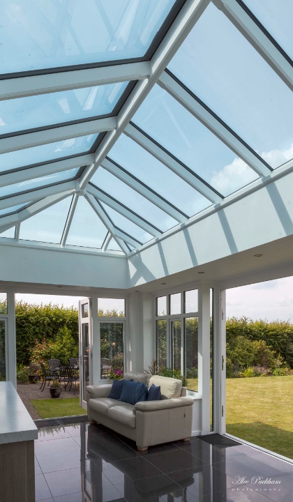 This is a bright, modern orangery with a large glass lantern roof that floods the space with natural light. The white-framed glazing blends with the crisp walls, while sleek black floor tiles reflect light for a spacious feel. A cream sofa with blue cushions adds comfort, and wide French doors open directly to a patio and garden seating area, creating a seamless indoor-outdoor connection.