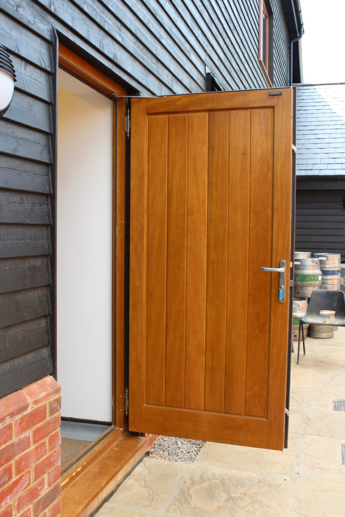 Country Hardwood solid oak front door with vertical panels, installed in timber clad house with brickwork detail.